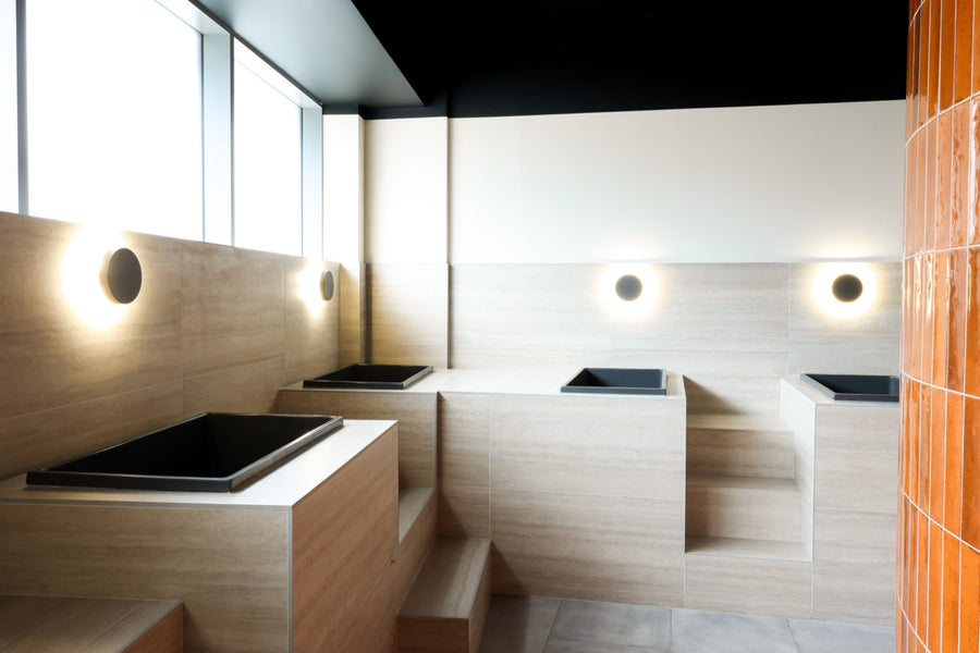 Matte-black plunge tubs set into travertine, beside the glass-brick drum